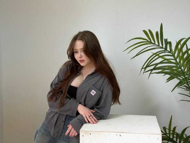 AlicaJames, avec de longs cheveux bruns, un t-shirt gris, un haut noir, un jean bleu, appuyée sur une boîte blanche, une plante verte à droite, regardant vers le bas, expression douce, lèvres légèrement entrouvertes, pose décontractée..