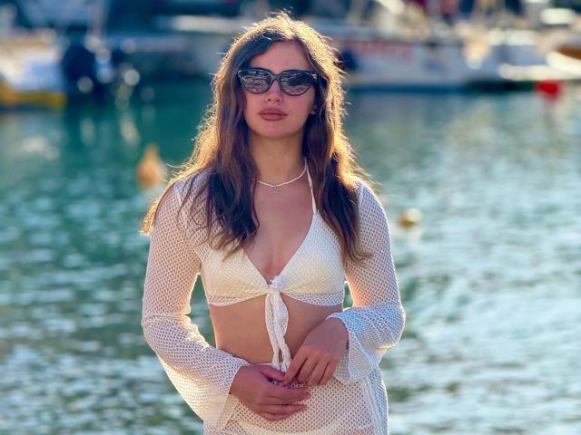 Dilana-hot with long brown hair, wearing a white mesh crop top and skirt, black sunglasses, and a necklace, standing by a waterway with boats in the background.