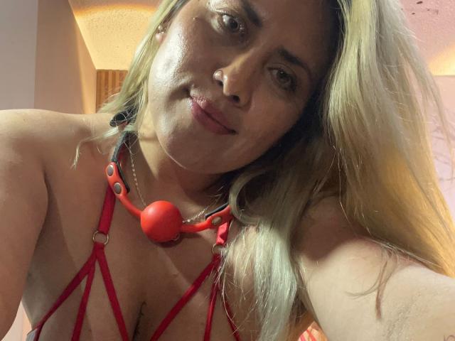 AlejaBlonde, com cabelo loiro, vestindo um top de lingerie vermelho com alças finas e uma boquinha vermelha, tem um leve sorriso e um piercing no nariz, em um quarto com pouca luz..
