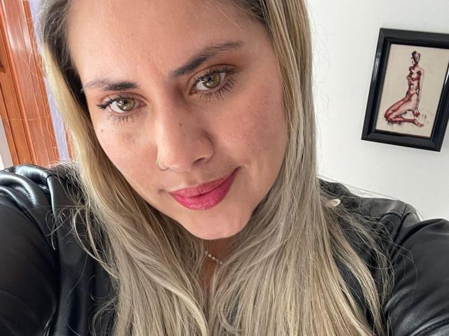 AlejaBlonde tiene cabello rubio, ojos verdes, lápiz labial rosa, una chaqueta de cuero negra, un piercing en la nariz, una ligera sonrisa y un dibujo enmarcado de una mujer desnuda en la pared blanca..