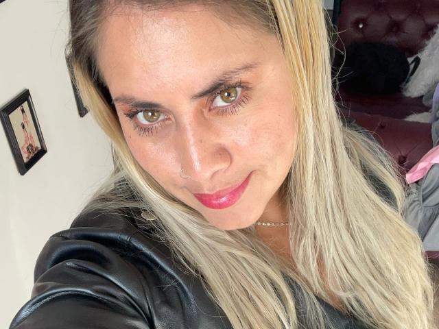 AlejaBlonde hat blondes Haar, braune Augen und einen Nasenring. Sie trägt eine schwarze Lederjacke und pinken Lippenstift und lächelt leicht. Ihre Brüste sind nicht sichtbar..