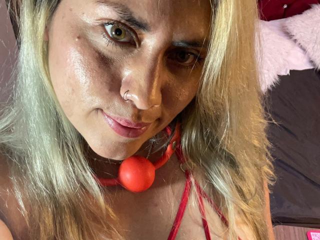 AlejaBlonde, con cabello rubio, aro en la nariz y un bozal rojo, tiene una piel brillante y ojos verdes, y está sonriendo ligeramente. Se puede ver su top rojo con tirantes..