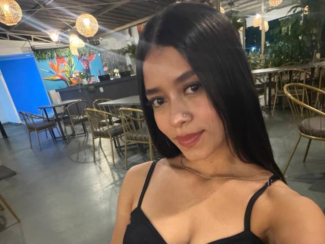 LaurentRay with long black hair, small nose ring, and black strap top, showing her tits in a dimly lit cafe with wicker chairs and hanging lights.