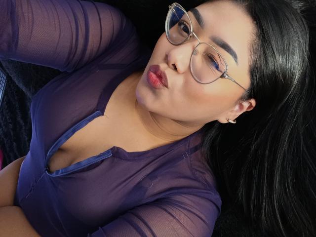 AlexaKiim with glasses, pouty lips, and dark hair, wearing a sheer purple top, showing her tits, lying on a black surface. Her eyes are half-open.