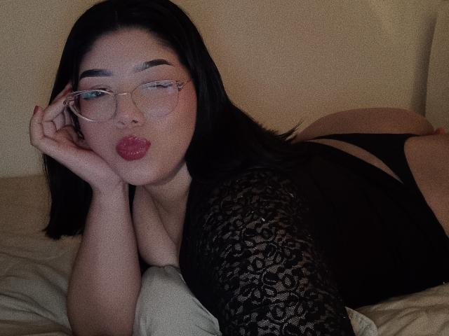 AlexaKiim with black lace top, glasses, and red lipstick, lying on bed, butt in black panties, hand on cheek, pouty lips, fair skin, long black hair, sexy.