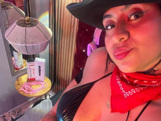 DiannaRosse with dark hair, red bandana, black hat, black bikini top, and pink lipstick, showing her boobs, sitting in a room with lamps and sex toys.