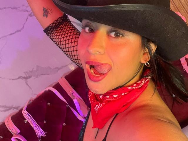 DiannaRosse with a black cowboy hat, tongue out, fishnet sleeve, red bandana, tattoo on arm, earrings, purple jacket, and pinkish lighting, showing off her sexy face.