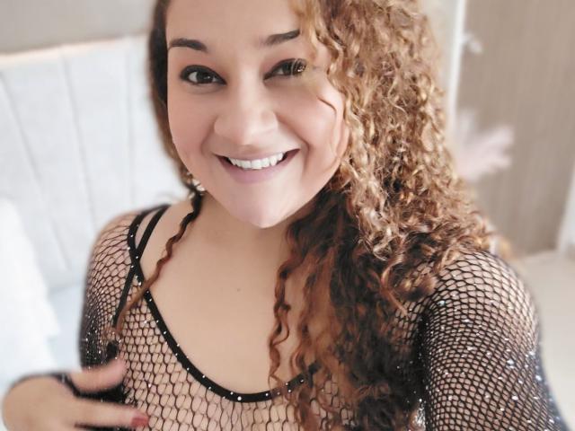 Natalia, met haar krullende bruine haar, glimlacht en draagt een zwarte top van netstof, waardoor haar kleine borsten zichtbaar zijn en een stukje van haar roze schaamlippen door het netwerk te zien is..