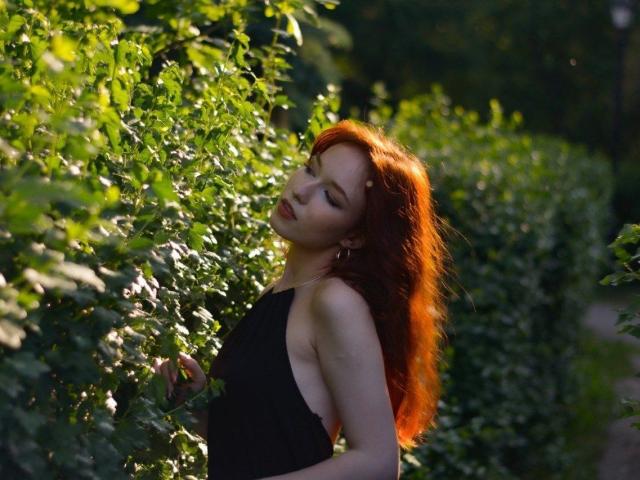 RandaFarrare with red hair, eyes closed, wearing a black dress, standing in greenery, her tits partially visible through the side, sunlight on her skin, relaxed, sensual.