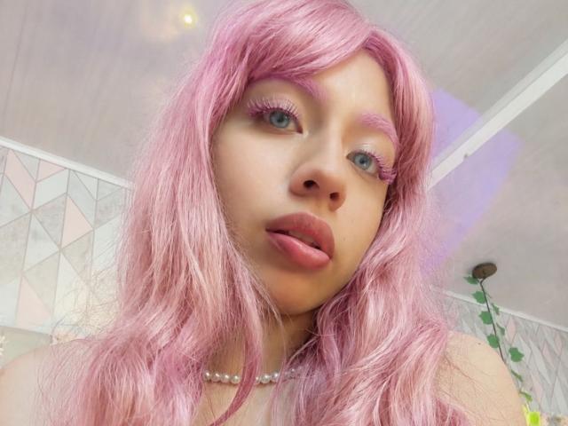 ZoeRedds has pink hair, blue eyes, and pink eyelashes. She's sticking out her pink tongue, wearing a pearl necklace. Her tits are partially visible. She's a cute bitch.