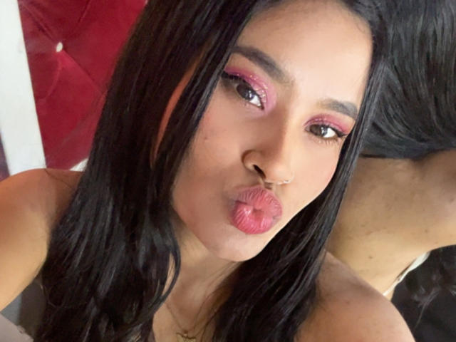 KarolPalacios, con cabello negro, sombra de ojos rosa, piercing en la nariz y labios rosados y fruncidos, haciendo una carita de beso. Su trasero es parcialmente visible en el fondo..