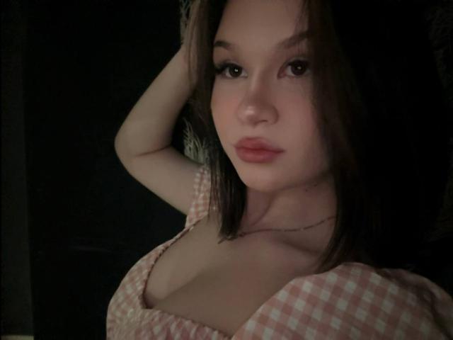 SaraJerome has small tits, a cute face, and brown hair in a checkered top, looking seductive with a hand in her hair, dark background, subtle lighting, soft expression.