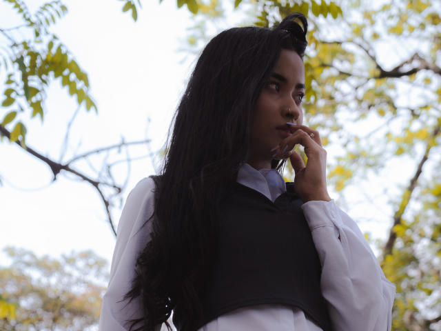 KarolPalacios with long black hair, white shirt, black vest, and finger near mouth, looking thoughtful. Yellow leaves in background. Soft light. Serious expression. Upper body shot.