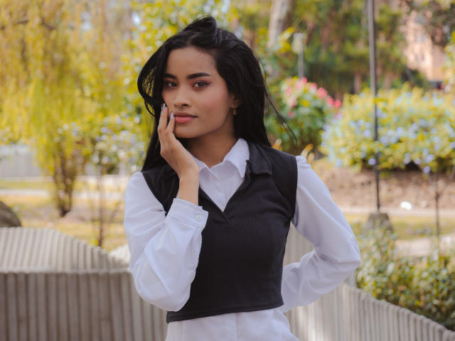 KarolPalacios, con cabello negro largo, camisa blanca y chaleco negro, tocándose la mejilla, con expresión pensativa, de pie en un jardín soleado, con las manos en las caderas, maquillaje sutil, expresión seria..