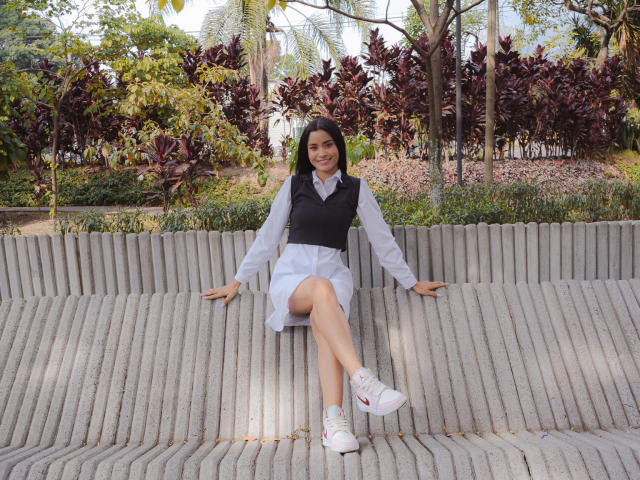 KarolPalacios, con chaleco negro, camisa blanca y falda, está sentada en un banco de madera, con las piernas cruzadas, zapatos blancos, sonriendo, cabello oscuro, en un parque soleado..