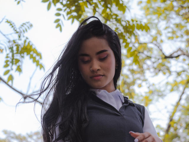 KarolPalacios with long black hair, nose ring, and pink eyeshadow, wearing a grey vest over a white shirt, eyes closed, looking down, arms crossed.