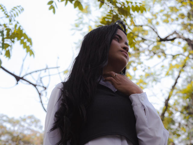 KarolPalacios with long black hair, wearing a white shirt and black vest, looking up, hand near neck, outdoors with green leaves and branches in background.