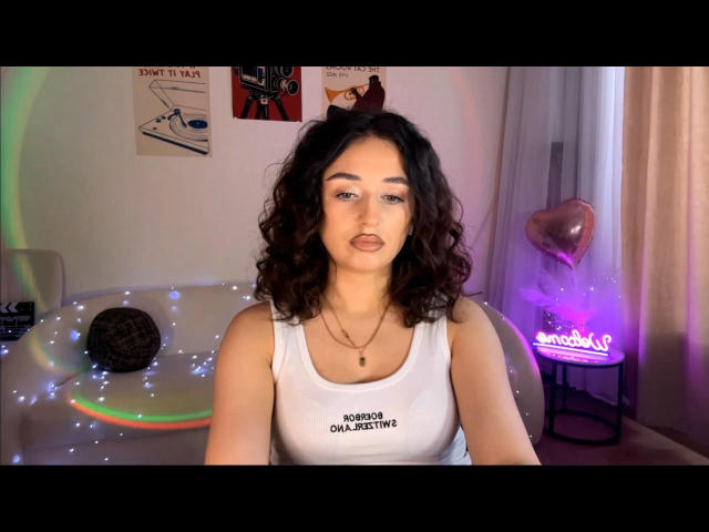 MyrtieGolds with curly dark hair, wearing a white tank top, has a neutral expression, sitting with a small round dark object on a table with fairy lights.