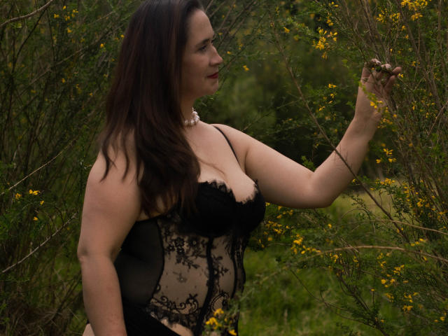 MatureTitsX69 con capelli scuri, indossa lingerie di pizzo nero, tocca fiori gialli, ha una collana di perle e guarda di lato, mostrando il seno..