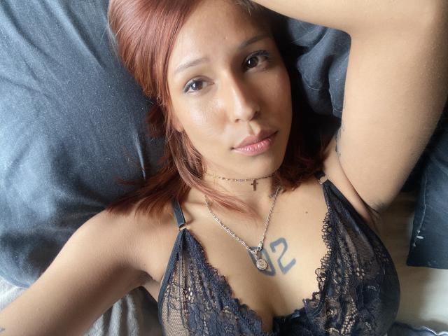 KiaraHernandezX with red hair, lying down, wearing black lace bra, small tattoo on chest, necklace, arm raised, smiling, brown eyes, light skin, bedroom setting, soft light.
