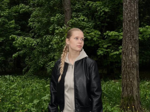 LillianaWhang has blonde braids, a black leather jacket, and a grey hoodie, standing in a forest with green trees and grass. Her ass is slightly visible.