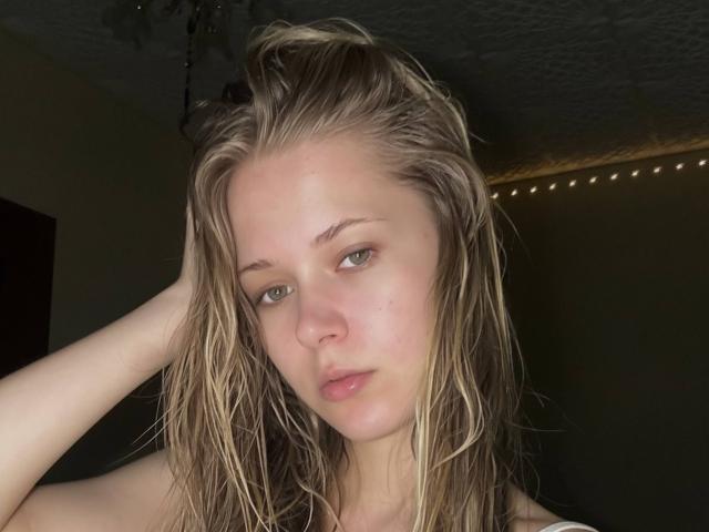 LillianaWhang has wet blonde hair, pale skin, and green eyes, looking slightly bored with her hand in her hair, showing her bare shoulders and subtle pink lips.