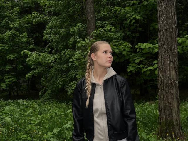LillianaWhang has blonde braided hair, wearing a black leather jacket over a beige shirt, standing in a green forest, looking up with a serious expression.