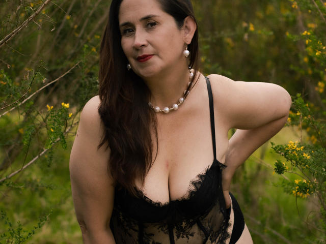 MatureTitsX69 met middelgrote borsten, gekleed in zwarte kant lingerie, met een parelketting en oorbellen, staand buiten, handen op de heupen, donkerbruin haar, rode lipstick en een lichte glimlach..