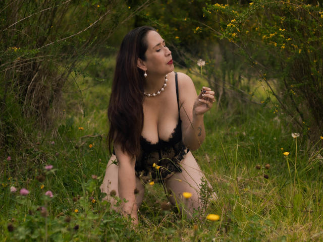 MatureTitsX69 with large boobs, kneeling in grass, wearing black lace lingerie, holding a dandelion, looking up, necklace, dark hair, green forest background, yellow flowers, serious expression.