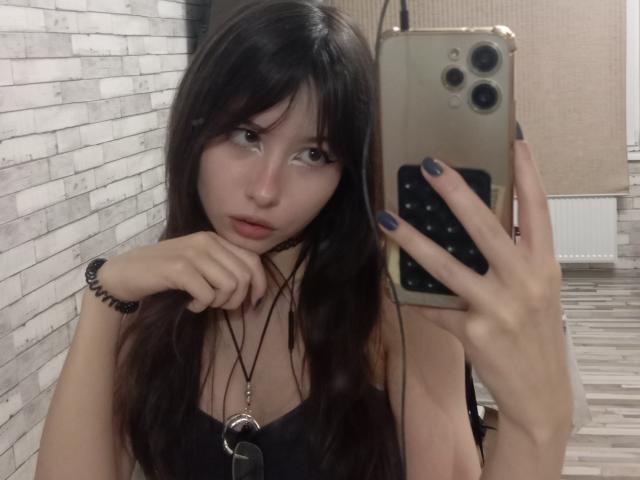 EileneSzala with long dark hair, holding phone, black nails, black dress, necklace, and wrist bracelet, looking at mirror, with a slightly pouty expression, in a selfie.