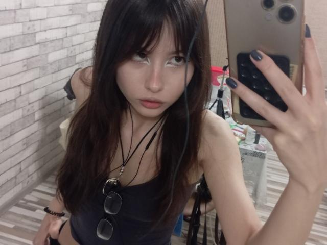 EileneSzala with long brown hair, wearing a black tank top, holding a phone, mirror selfie, blue-gray nail polish, black necklace, small tits, pale skin, and a serious expression.