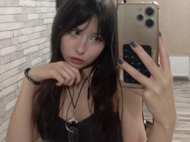 EileneSzala with long dark hair, holding a phone, wearing a black top, necklace with a ring, and black nails, looking in a mirror, with a slightly pouty expression.