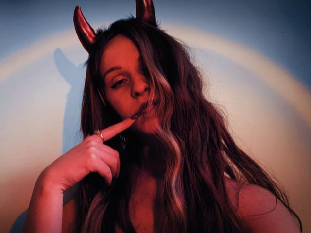 FlyFreedom with long wavy brown hair, red devil horns, and a finger seductively touching her lips. She's looking sexy with a slight smile and a shadow on the wall.