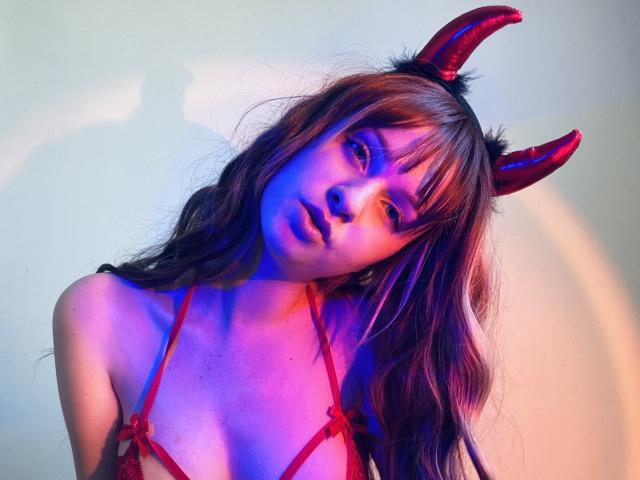 FlyFreedom with red devil horns, purple-blue lighting, and long brown hair, wearing a red lace bra, looking slightly down with a pouty expression. Her tits are visible.