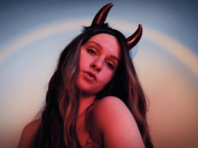FlyFreedom with long brown hair, wearing shiny red devil horns, looking at the camera with a slight pout, pinkish-red lighting on her face and upper body.