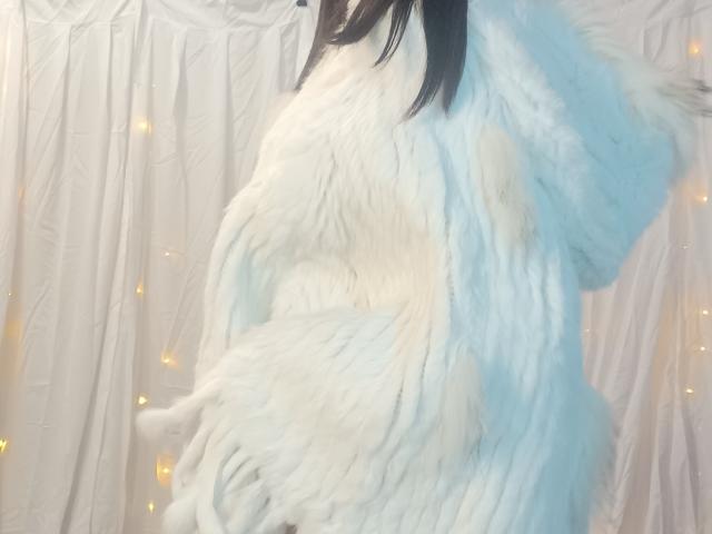OrianaLaFrancaise in a fluffy white and light blue fur coat, her dark hair falling to the side, with soft yellow fairy lights in the background.