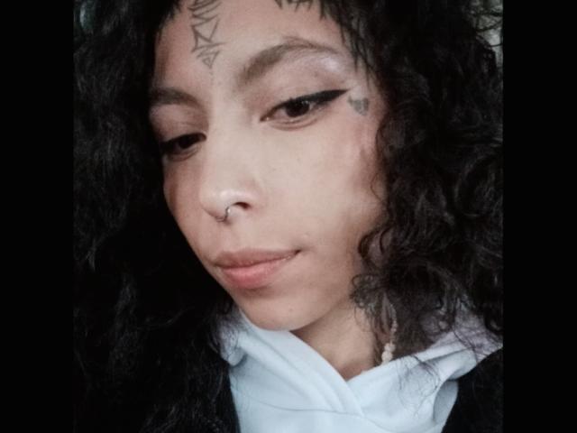 LaurieRzz a les cheveux noirs et bouclés, un piercing au nez et un eye-liner noir avec un effet "ailes de papillon". Elle a des tatouages sur le front et près de l'œil. Elle porte un sweat à capuche blanc..