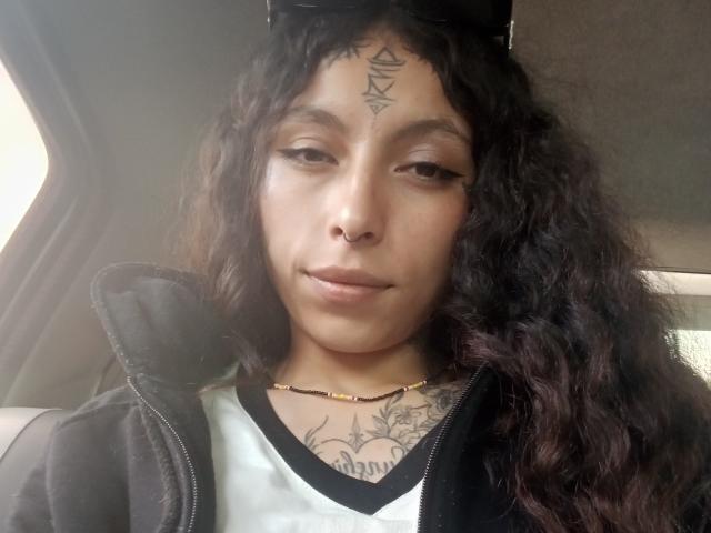 LaurieRzz mit lockigem, dunklem Haar, Nasenring, schwarzem Tattoo über der Stirn und einem Brusttattoo, trägt eine schwarze Jacke und ein weißes Hemd und sitzt in einem Auto, leicht lächelnd..
