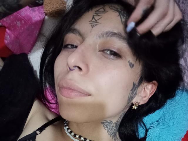 LaurieRzz, com cabelo preto, piercing no nariz, tatuagens e brincos, deitada, com a mão no cabelo, usando um colar, lábios levemente curvados, unhas azuis, sorriso suave e um pequeno piercing no nariz..