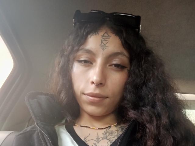LaurieRzz ha i capelli ricci e scuri, un piercing al naso e un tatuaggio a forma di triangolo sulla fronte. Indossa una giacca nera con gli occhiali da sole sopra. Si vedono dei tatuaggi sul petto..