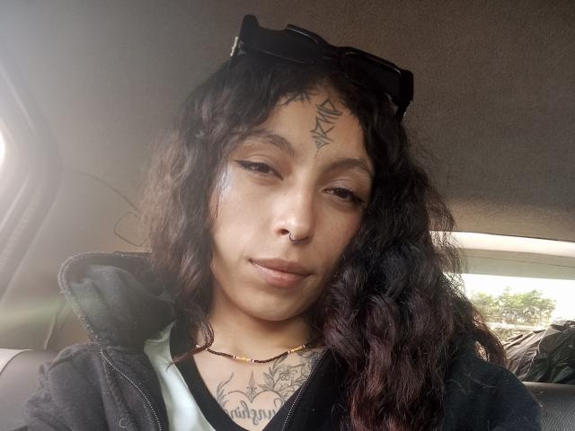 LaurieRzz, con cabello negro rizado, cejas oscuras, piercing en la nariz, tatuajes en la frente y el pecho, chaqueta negra, gafas de sol en la cabeza, una sonrisa sutil, interior de un coche, luz solar..