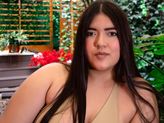 ScarlettVex has long dark brown hair, a fair skin tone, and wears a beige halter top, showing her small tits, with a confident, slightly smug expression on her face.