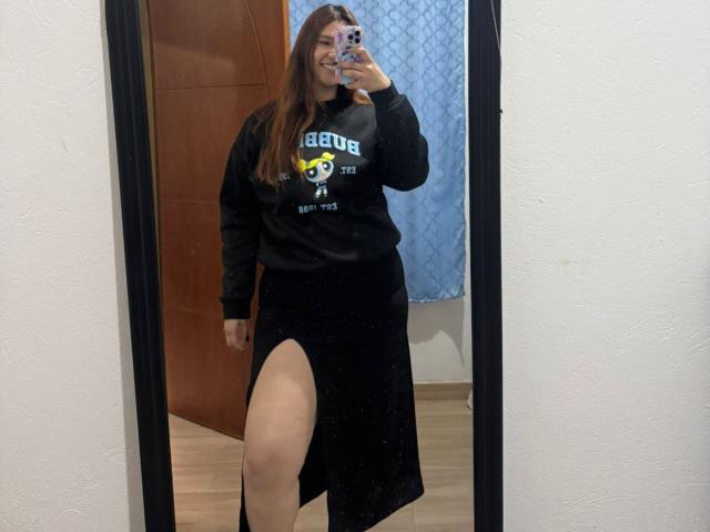 SofiaVelvet ha lunghi capelli castani, una felpa nera Adidas e una gonna nera con una spacca alta che mostra la sua coscia. Sta sorridendo e sta scattando un selfie allo specchio..