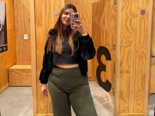 SofiaVelvet has long brown hair, a black velvet jacket, grey crop top, and green pants, taking a mirror selfie with her phone in a wooden changing room.