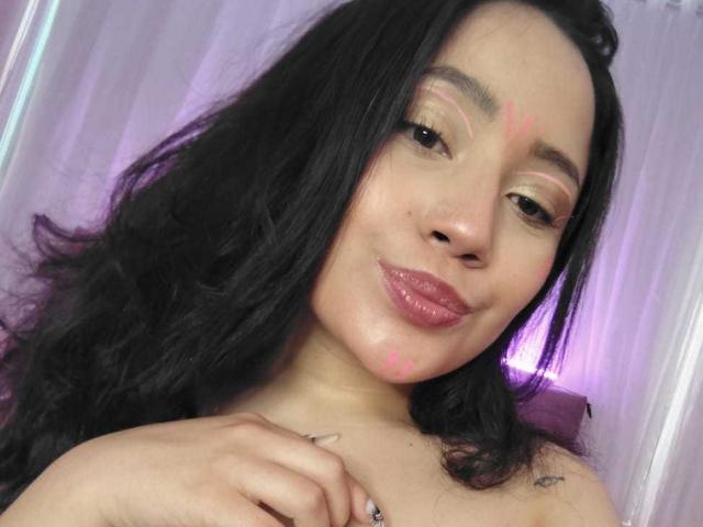 AlessaMora with dark, wavy hair, pink lipstick, and subtle eye makeup, smiling, has pink face markings, and her hand rests on her shoulder, showing a small tattoo.