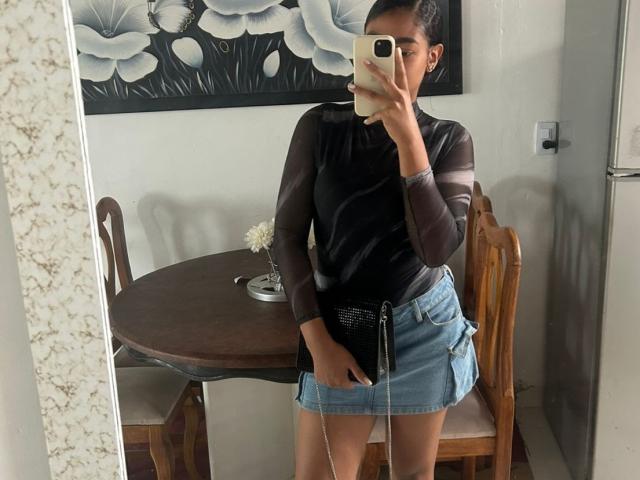 NatalyBlonder trägt ein durchsichtiges schwarzes Oberteil, einen blauen Jeansrock und macht ein Spiegel-Selfie. Sie hält ihr Handy, wobei ihre Hand im Rock zu sehen ist, und das Spiegelbild spiegelt sich darin..