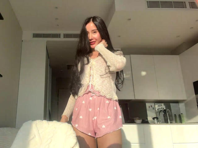 Kandace, com cabelos longos e pretos, vestindo um cardigã creme e shorts rosa com estampas de corações, segurando um cobertor branco, com a luz do sol em seu rosto, em pé em uma cozinha moderna..