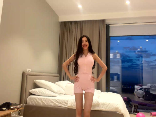 KandaceMoles está de pie en un dormitorio, vestida con una camiseta rosa sin mangas y pantalones cortos, con las manos en las caderas, cabello castaño largo, sonriendo, una cama blanca con almohadas detrás de ella, se ve un cielo nocturno..