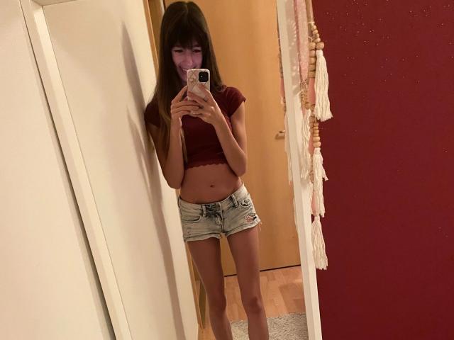 LaylavonHohensee – heiß, mit langen braunen Haaren, einem roten Crop-Top und Jeansshorts, macht ein Selfie vor einem Spiegel und zeigt dabei ihren nackten Bauch und ihre Beine, während sie ein rosa Handy hält..
