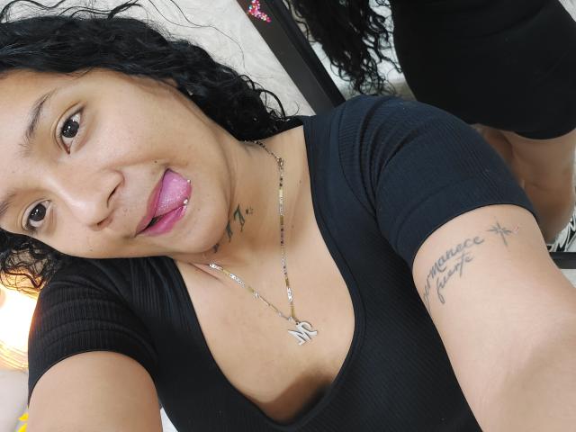 Kimberly, avec ses cheveux noirs bouclés, sa langue rose dépassant, a un tatouage sur son bras droit et porte un t-shirt noir côtelé qui révèle ses seins..